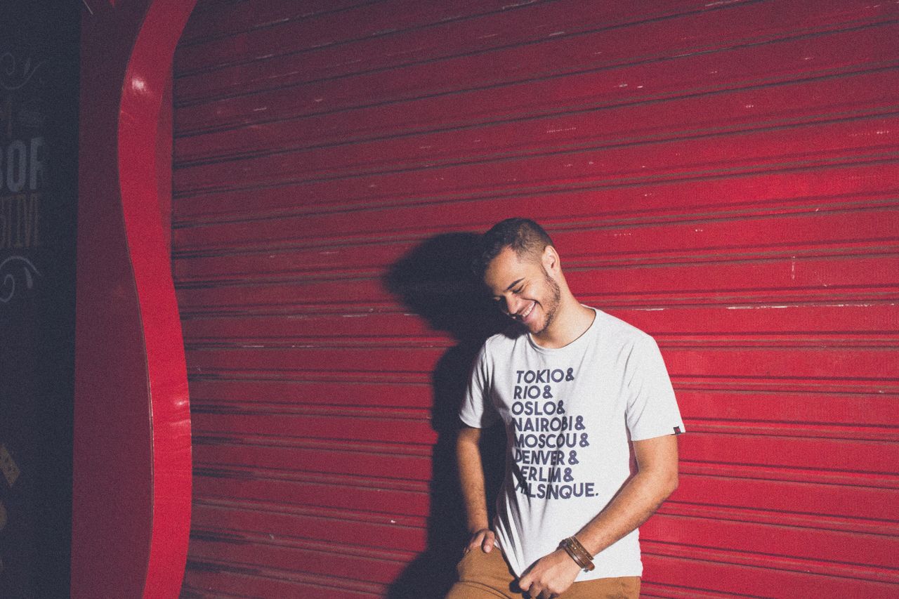 Casually dressed young man posing playfully against a vibrant red steel shutter with text t-shirt design.