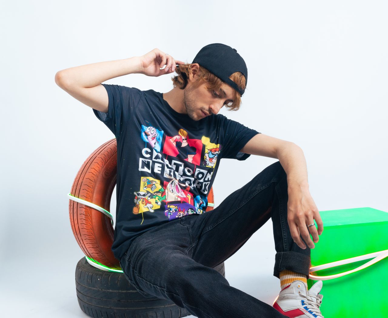 A young man wearing a graphic cartoon T-shirt sits on tires in a stylish pose.
