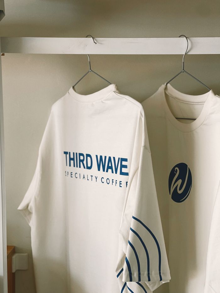 White t-shirts hanging with 'Third Wave Specialty Coffee' branding. Minimalist design.